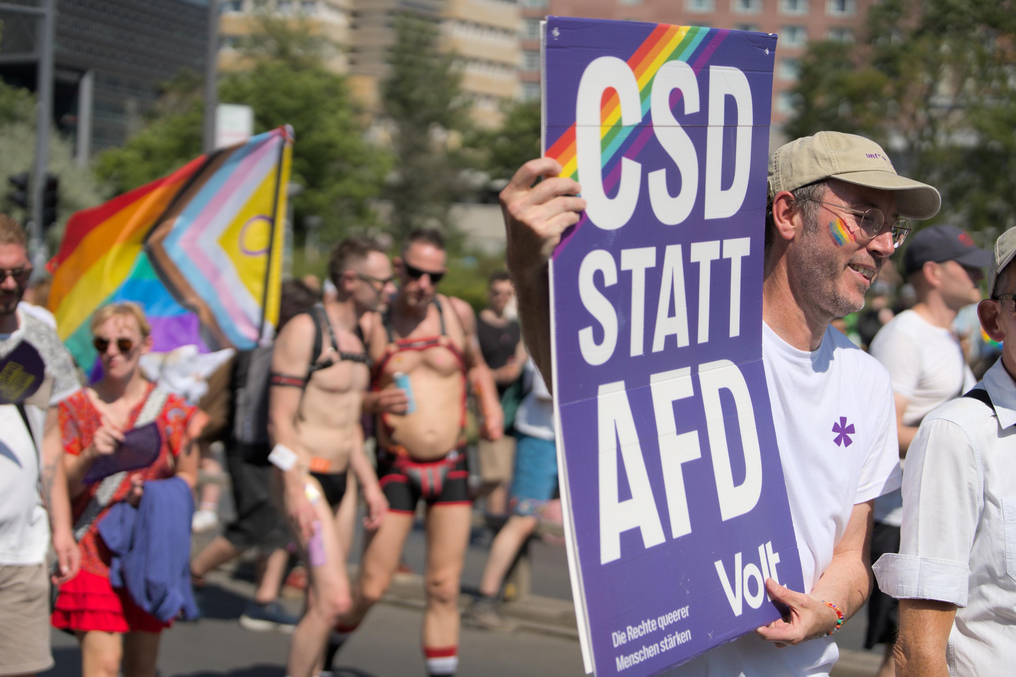 A person holding a Volt signs saying “CSD STATT AFD”. We can see a couple in the back with a rainbow flag.
